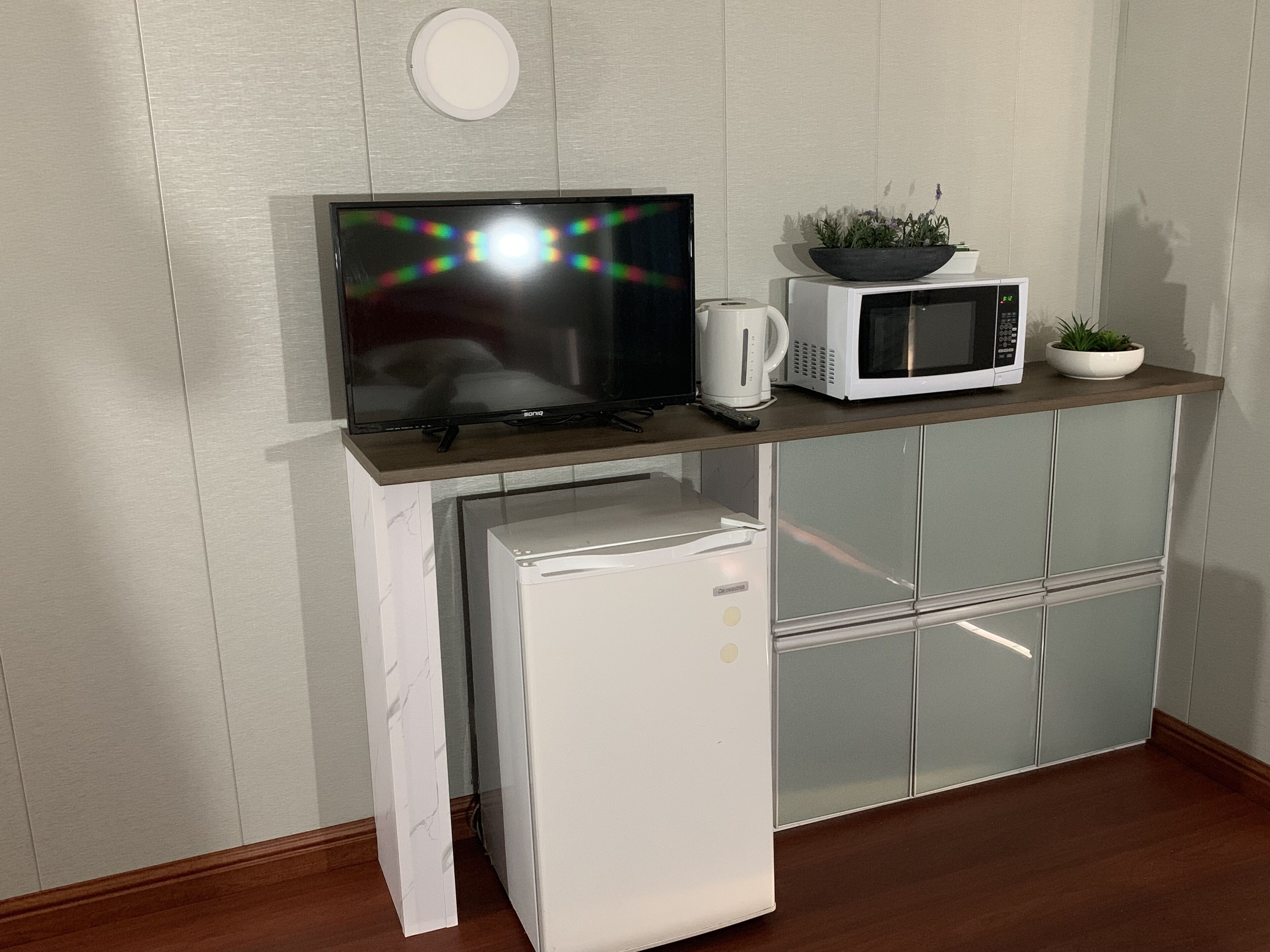 Eine kompakte Küchenzeile mit Flachbildfernseher, Mikrowelle, Kaffeemaschine und kleinem Kühlschrank.