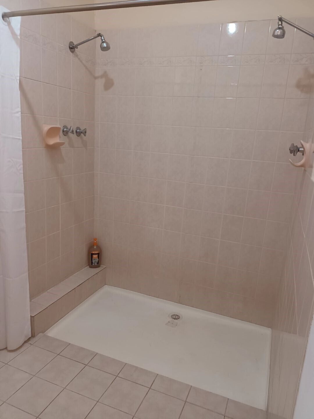 A walk-in shower with beige tiles, a handheld showerhead, a handheld showerhead, and a bottle of shampoo on the floor.