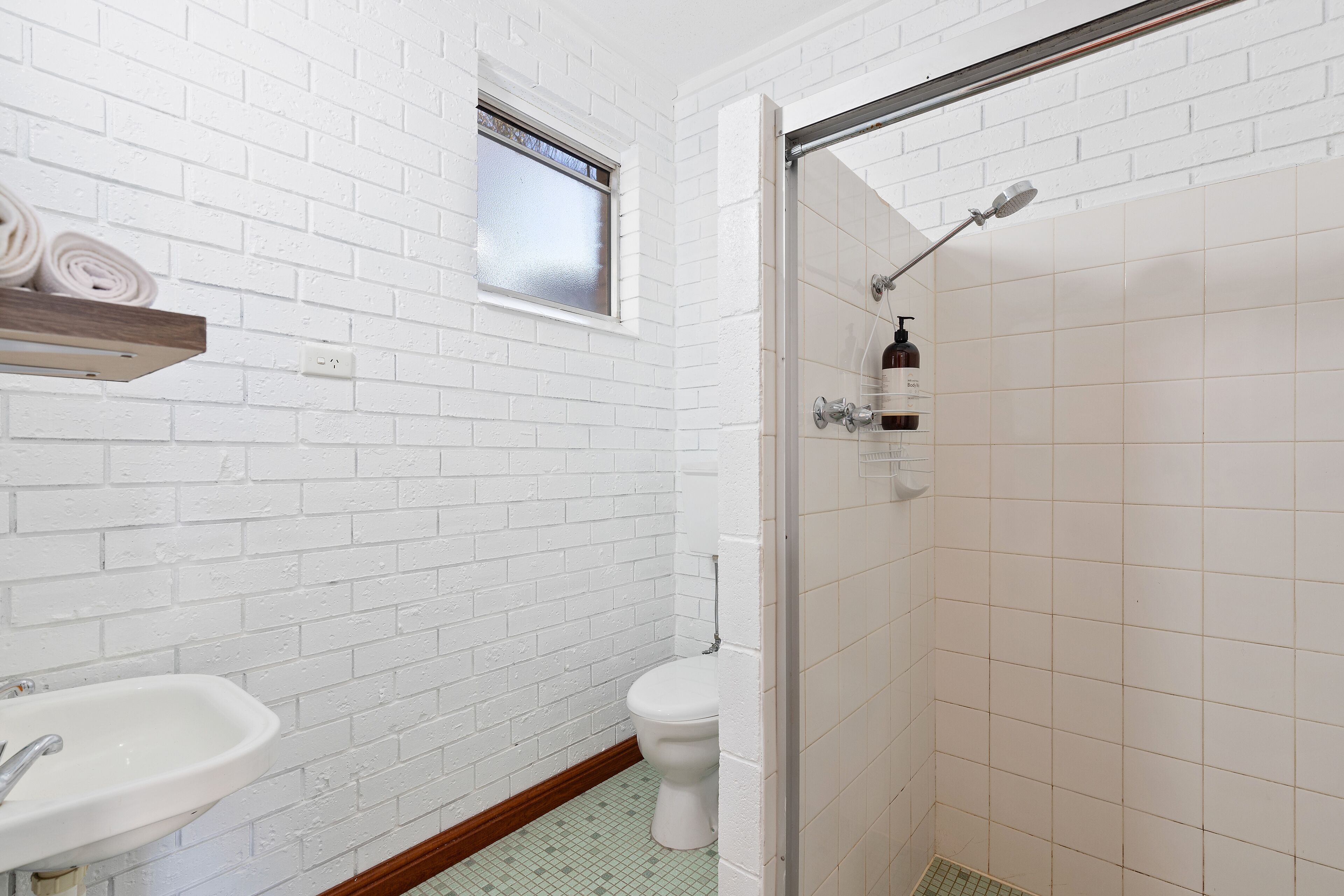 A bathroom with white brick walls, a white toilet, a sink with a wooden shelf above it, and a shower area.