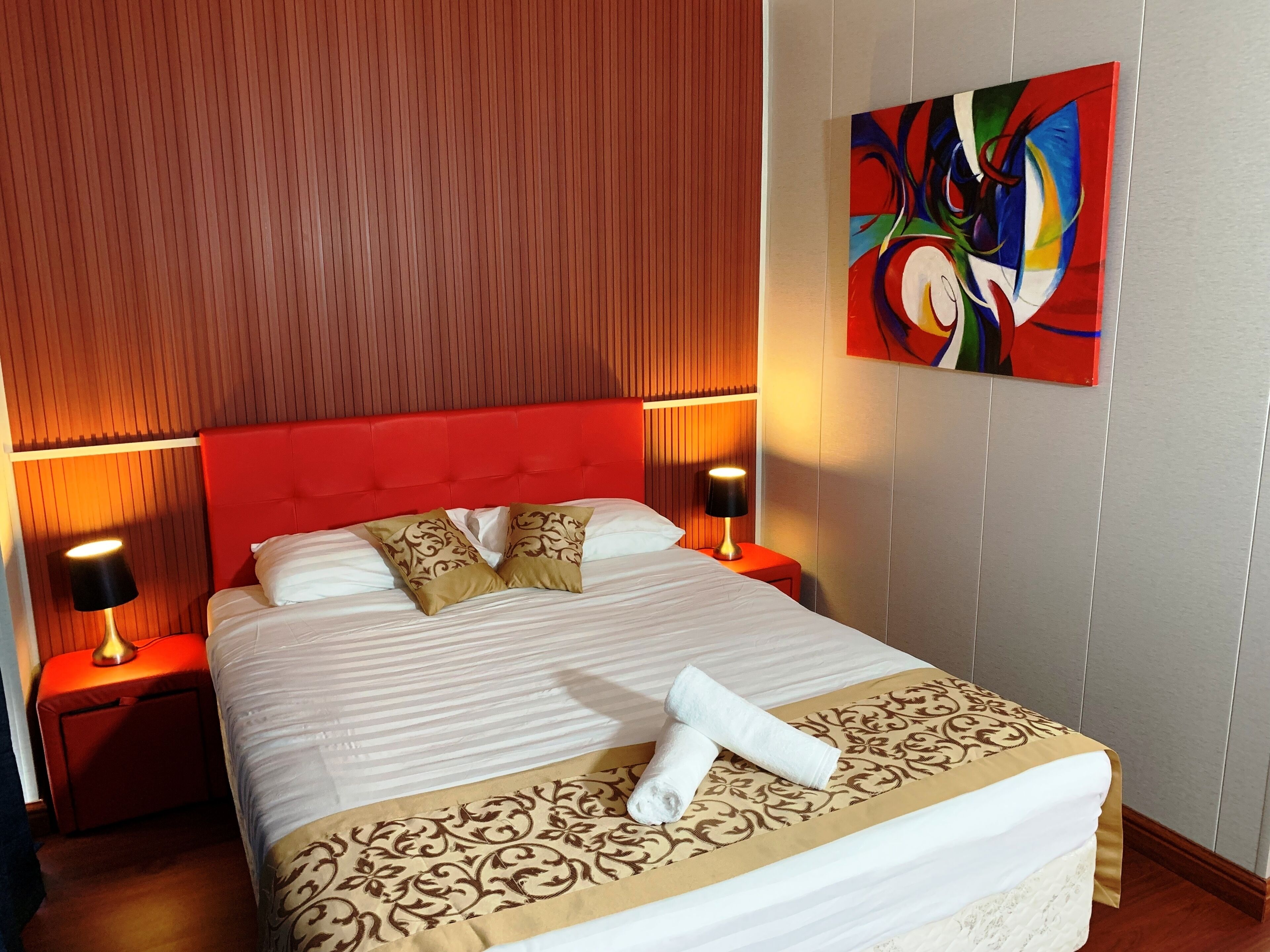Quarto de hotel com cabeceira vermelha, duas luminárias de cabeceira, um quadro grande e uma cama bem arrumada com colcha e almofadas estampadas.