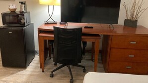 A compact hotel room with a wooden desk, a large flat-screen TV, a small refrigerator, a microwave, a lamp, and a potted plant. - The Hotel Orange (Orange)