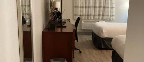 Une chambre d’hôtel avec un plancher en bois, un bureau avec une chaise, un lit, une fenêtre avec des rideaux et un radiateur.