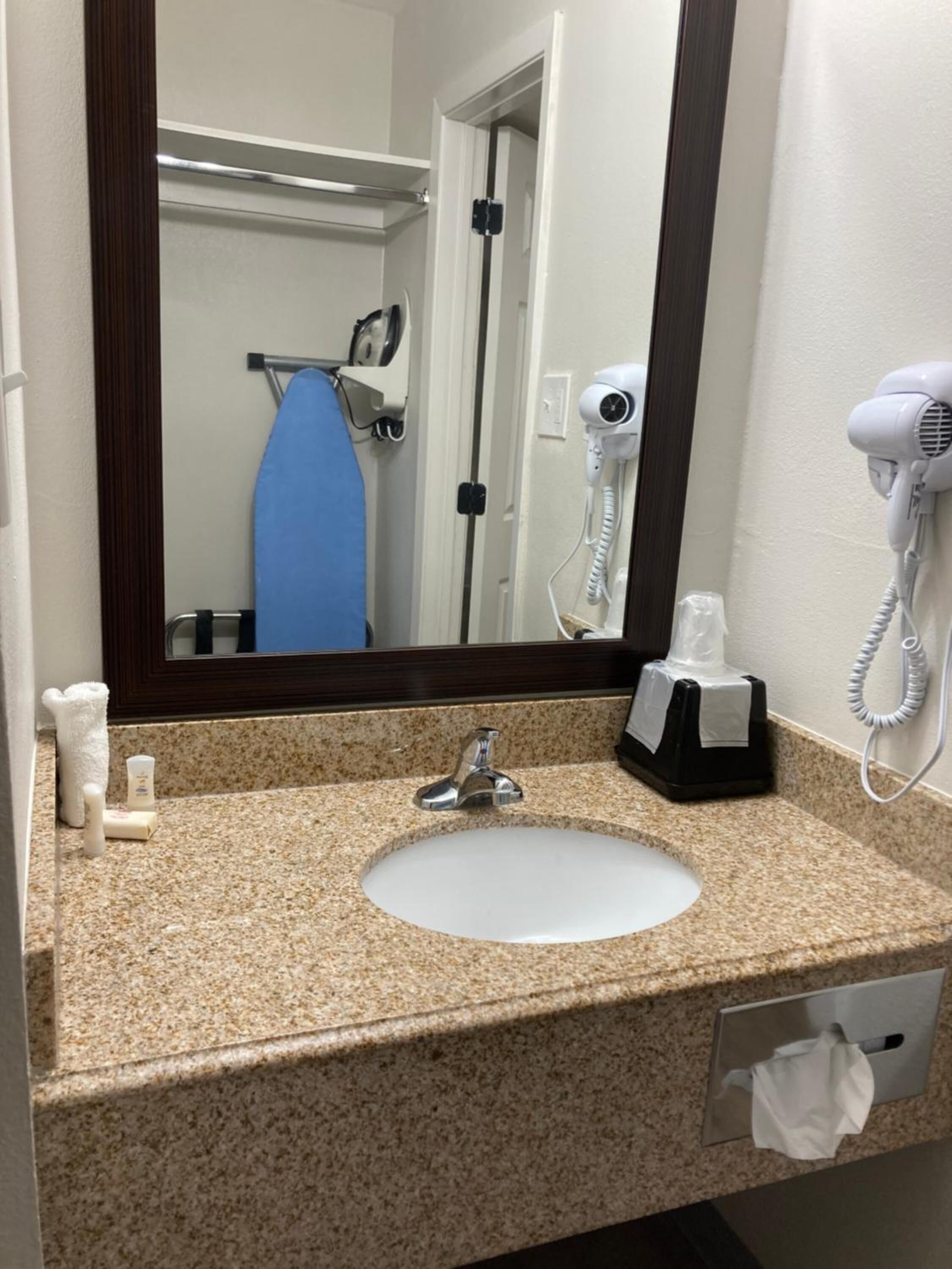 A bathroom with a granite countertop, a white sink, a mirror, a hairdryer, and a towel dispenser.