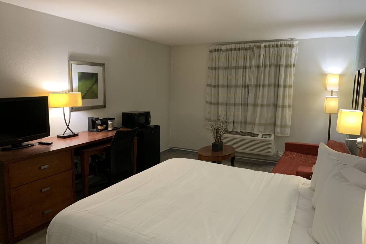 Ein Hotelzimmer mit einem Bett, einem Schreibtisch mit Fernseher und Lampe, einem Sessel, einem kleinen Tisch ebenfalls mit Lampe und einem Fenster mit Vorhängen.