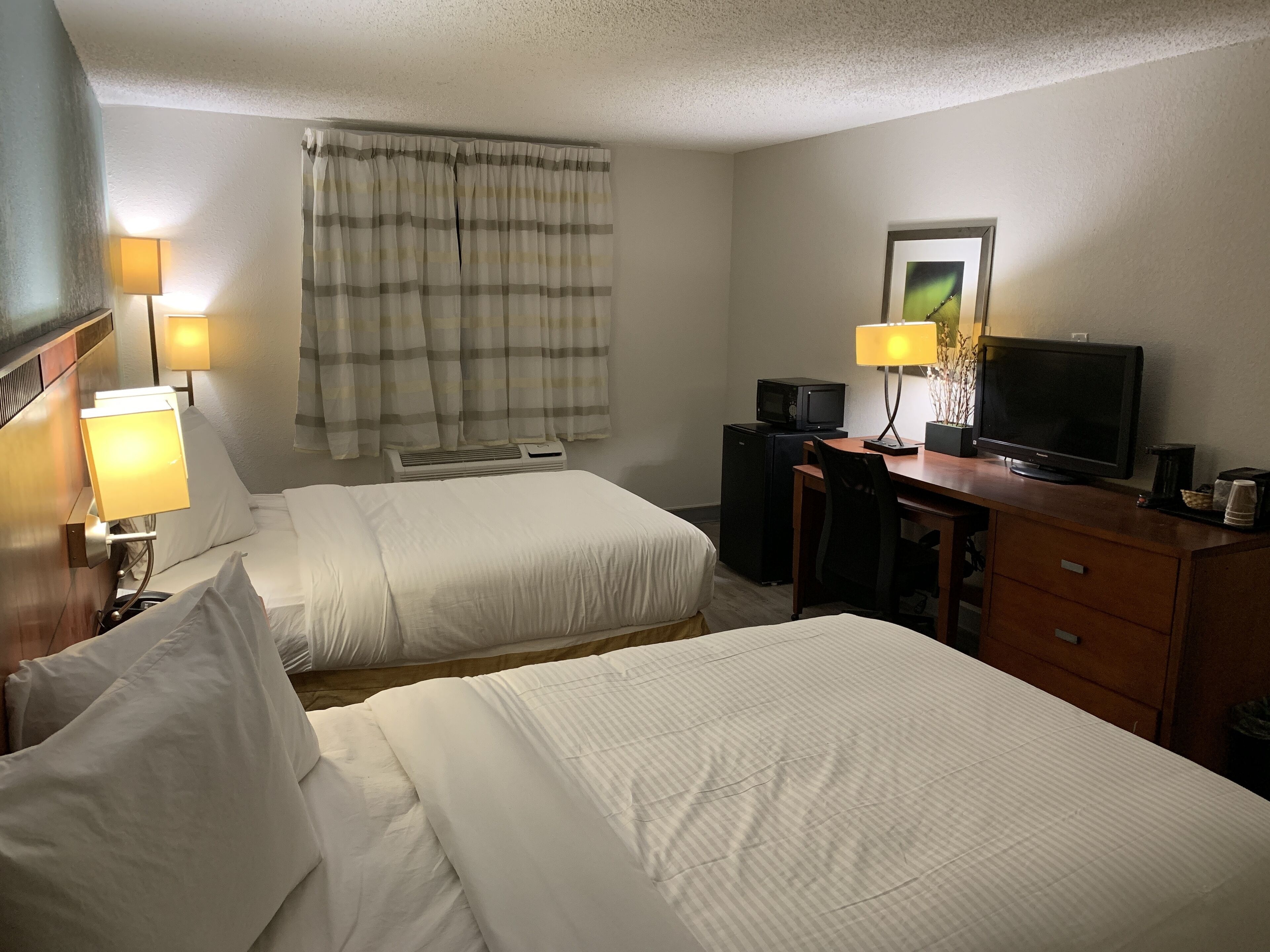 Ein Hotelzimmer mit zwei Betten, einem Schreibtisch mit Fernseher, einer Schreibtischlampe und einem Fenster mit Vorhängen.