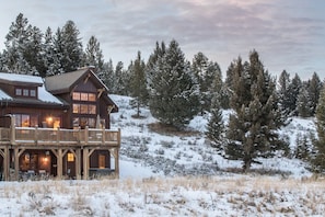 A large log cabin with a balcony, surrounded by snow-covered trees and a hilly landscape.