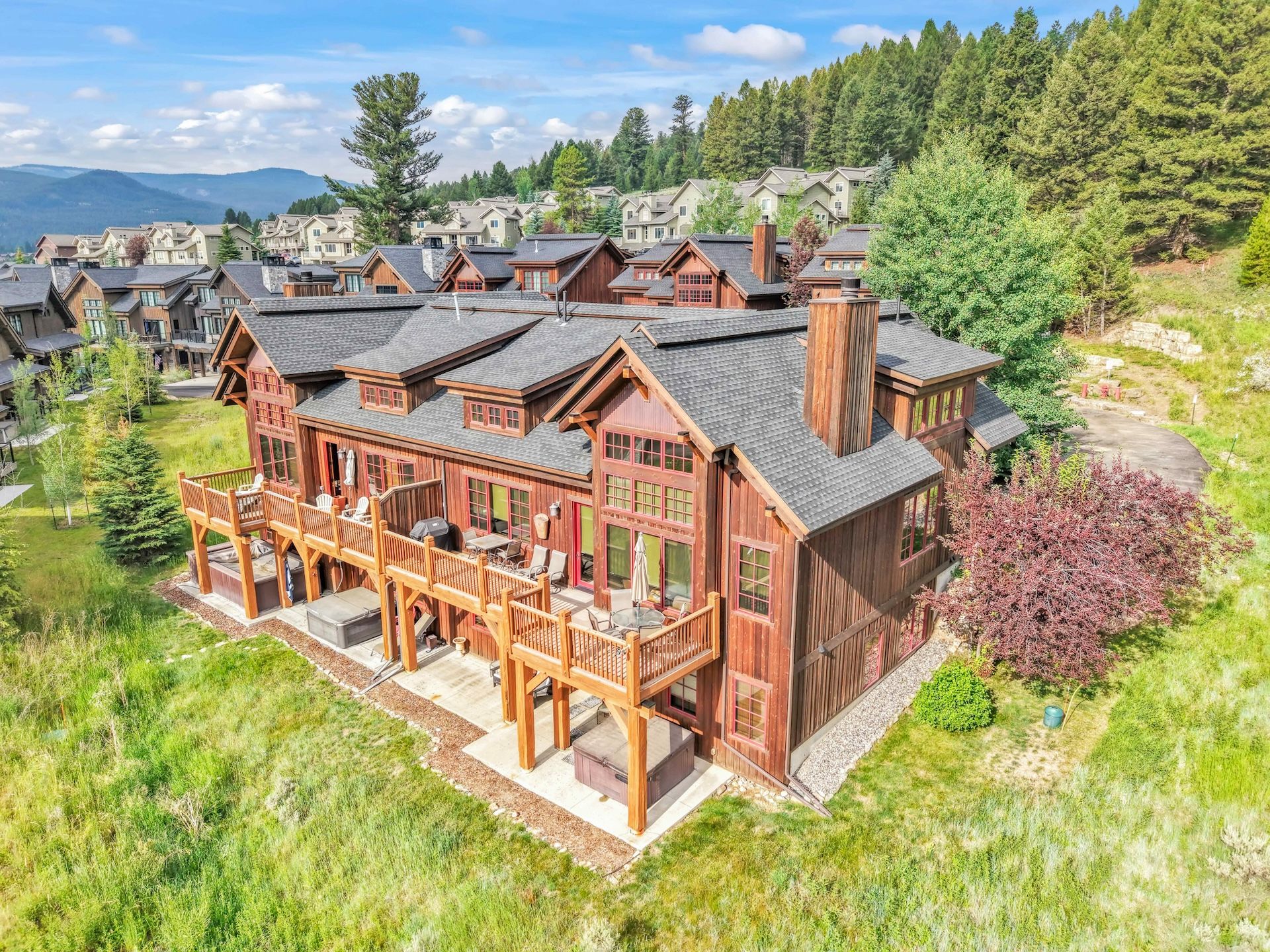 A large wooden house with a deck and multiple windows, surrounded by a lush green landscape and other houses.