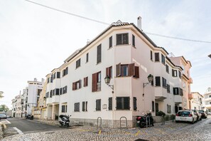 Un immeuble à plusieurs étages, doté d’une façade blanche et de volets bruns, devant lequel se trouve une rue pavée.