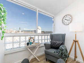 Une pièce avec vue sur la ville, un fauteuil gris, une petite table avec un plateau, une horloge et une lampe.