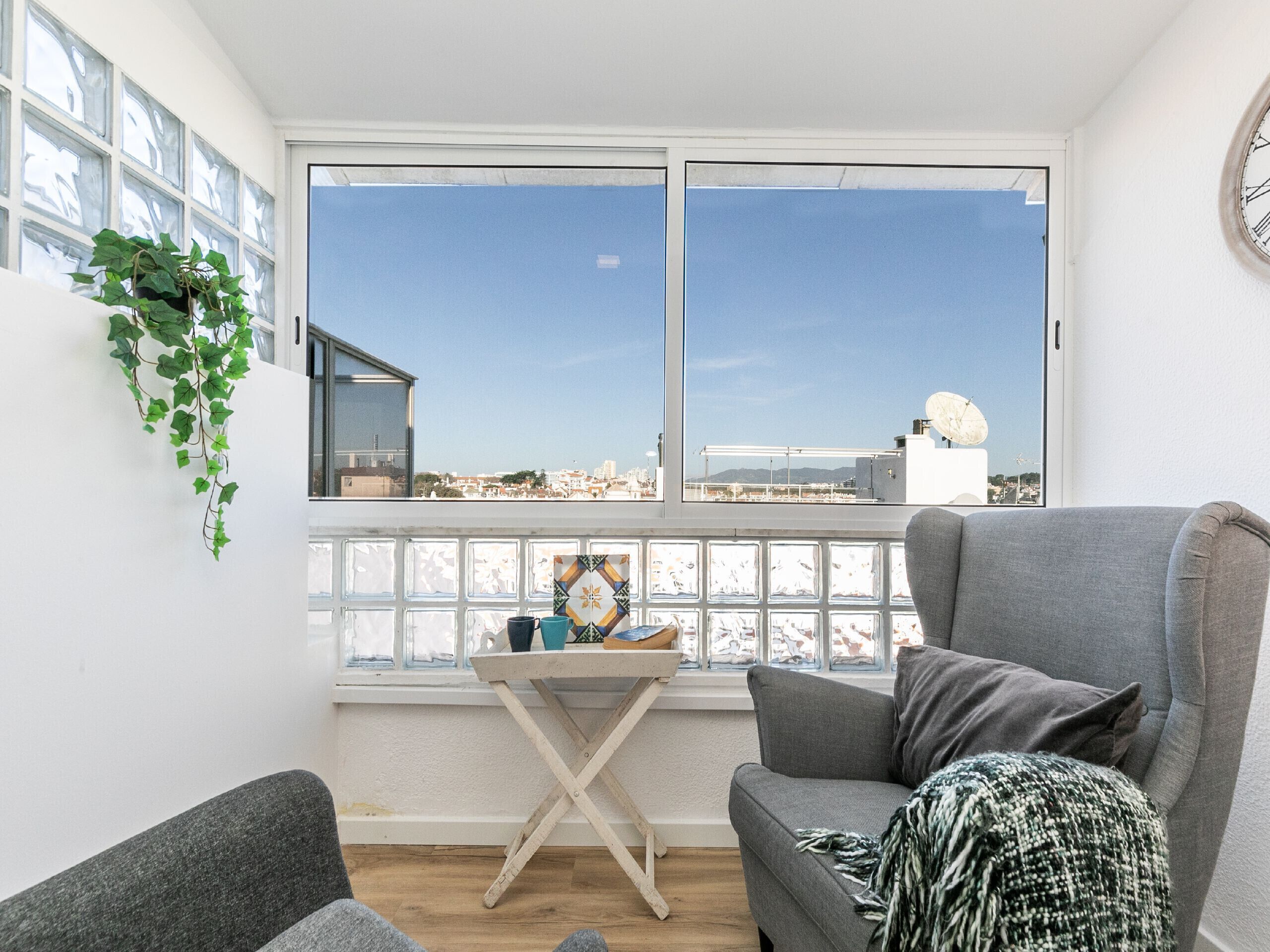 Un salon avec un canapé gris, une petite table en bois et une vue sur la ville grâce à de grandes fenêtres.