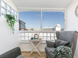 A modern living room with a grey sofa, a small wooden table, and a view of a cityscape through large windows.