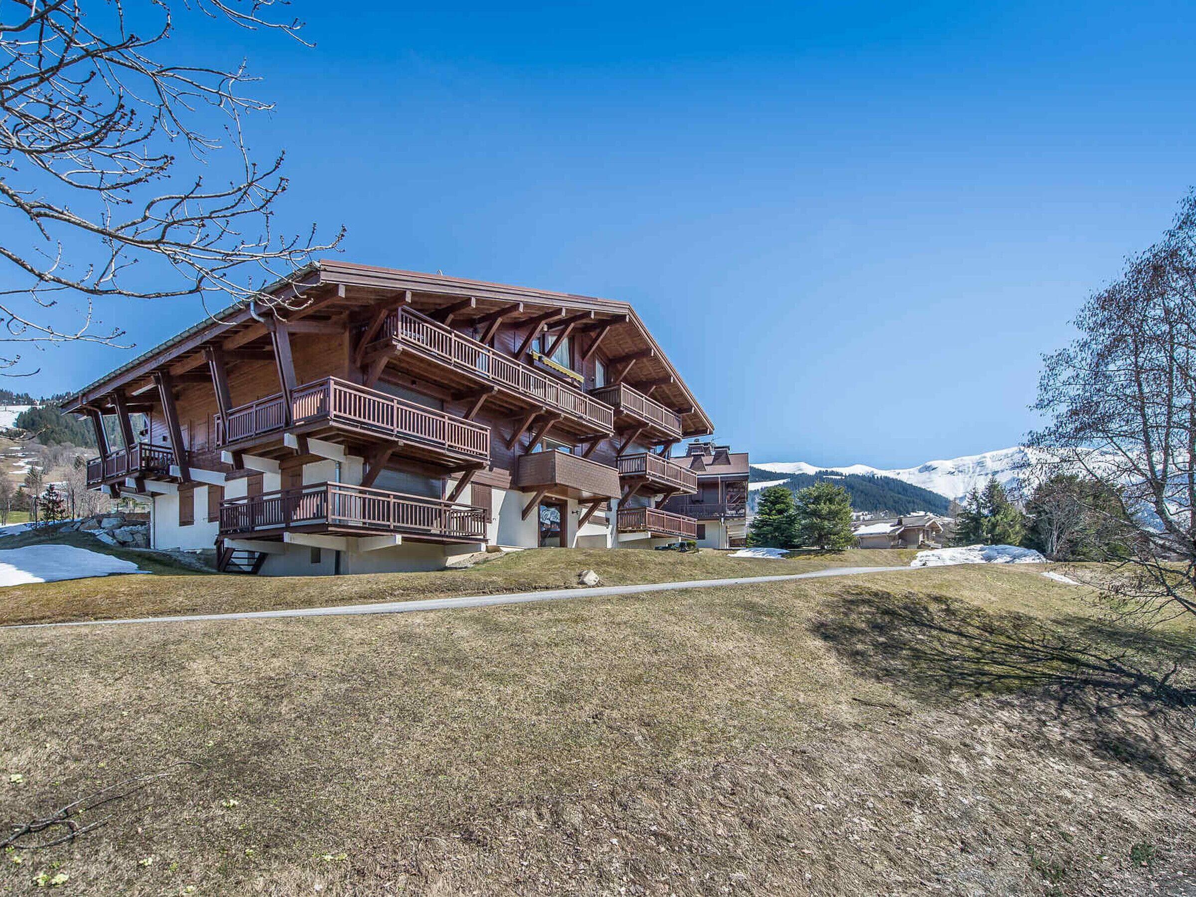 A multi-story wooden chalet with balconies, set in a mountainous area with snow-capped peaks in the background.