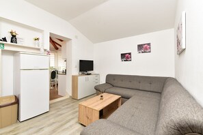Un salon moderne avec un canapé gris, une table basse en bois, un téléviseur et un coin cuisine équipé d’un réfrigérateur et de placards.