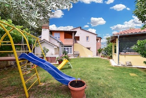 Zona residencial con un parque infantil, que incluye un tobogán y una estructura para escalar, un césped cuidado y casas de diferentes estilos arquitectónicos.