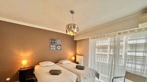A hotel room with two beds, a desk, a chair, a radiator, and a window with curtains. - Branly 2,2 bedrooms and 1 parking (Cannes)
