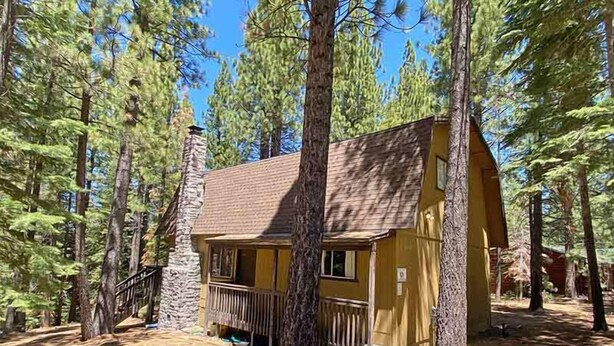 A cabin with a stone chimney, a wooden porch, and a clear view of the surrounding forest.