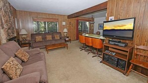 A cozy living room with a brown sofa, a dining table with orange chairs, and a flat-screen TV on a wooden stand.