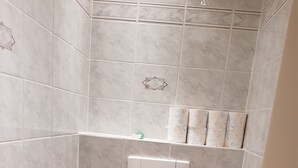 Een kleine betegelde badkamer met een toilet, een klein prullenbakje en een plank met toiletartikelen.