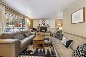 Un salon chaleureux avec une cheminée en pierre, deux canapés, une table basse et une grande fenêtre donnant sur un paysage enneigé.