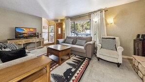 Olohuone, jossa on sohva, nojatuoli, sohvapöytä, televisio ja ikkunasta avautuva näkymä ulos.