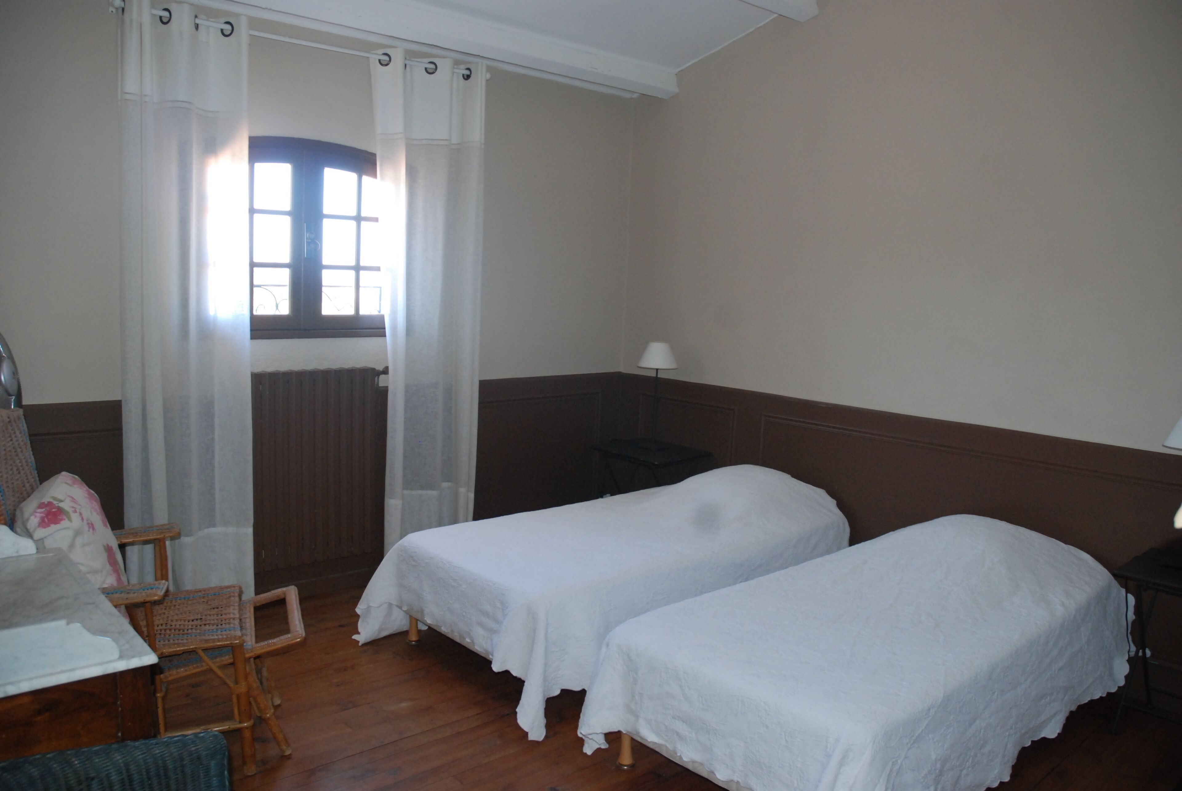 Una habitación con dos camas individuales, una silla, una mesa, una lámpara y un ventanal con cortinas.