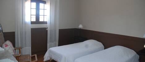 Una habitación con dos camas individuales, una silla, una mesa, una lámpara y un ventanal con cortinas.