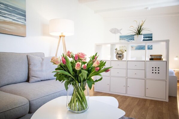 A living room with a grey sofa, a white coffee table, and a vase of pink flowers. There is a white cabinet with decorative items and a lamp. - Small hideaway<br>- A very special domicile in the middle of Friedrichstrasse -<br>The large double bed (180x200, mattress size approx. 1.92 m x 90 cm) is located in the modern living room, separated by a room divider. The flat screen TV. (Westerland)