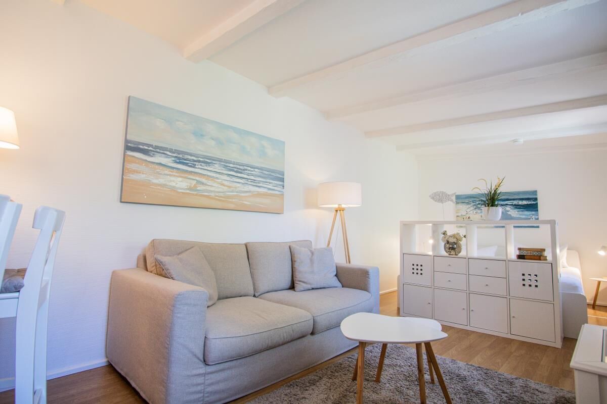 Ein Wohnzimmer mit einer Couch, einem kleinen Tisch und einem Schrank. Zwei Lampen und ein Gemälde sind an der Wand angebracht.