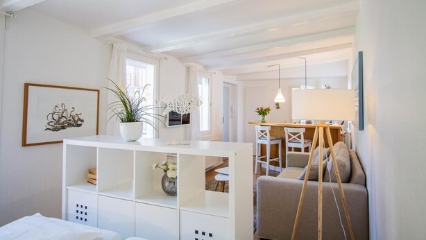 A modern, well-lit room with a white console table, a sofa, and a dining area with a wooden table and chairs. - Small hideaway<br>- A very special domicile in the middle of Friedrichstrasse -<br>The large double bed (180x200, mattress size approx. 1.92 m x 90 cm) is located in the modern living room, separated by a room divider. The flat screen TV. (Westerland)