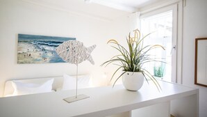 A white room with a painting of a beach scene, a white chair, and a potted plant on a white table. - Small hideaway<br>- A very special domicile in the middle of Friedrichstrasse -<br>The large double bed (180x200, mattress size approx. 1.92 m x 90 cm) is located in the modern living room, separated by a room divider. The flat screen TV. (Westerland)
