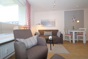 A living room with a sofa, armchair, and a small table with a lamp. There is a window with curtains, a painting on the wall, and a table with chairs. - The 2-room apartment ``Ankerplatz`` in the house Sola Bona is located on the 1st floor in a central location on Friedrichstraße in Westerland. The beach and pedestrian areas are only a few meters away. The apartment is for 2 people on around... (Westerland)