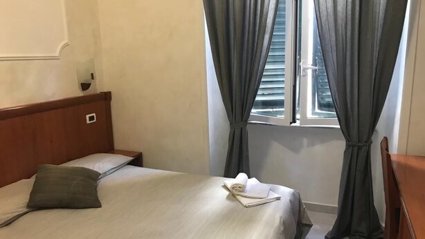 A hotel room with a bed, a wooden headboard, a window with curtains, and a small table with a chair. - Les Artes (Rome)