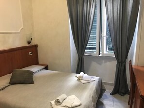 Habitació d'hotel amb un llit, un panell de capçalera de fusta, una finestra amb cortines i una taula petita amb una cadira.