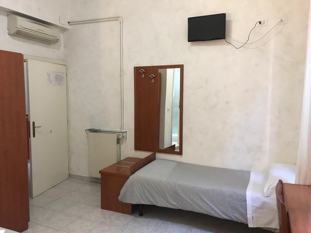 A small, single-bed room with a wooden wardrobe, a mirror, and a TV mounted on the wall.