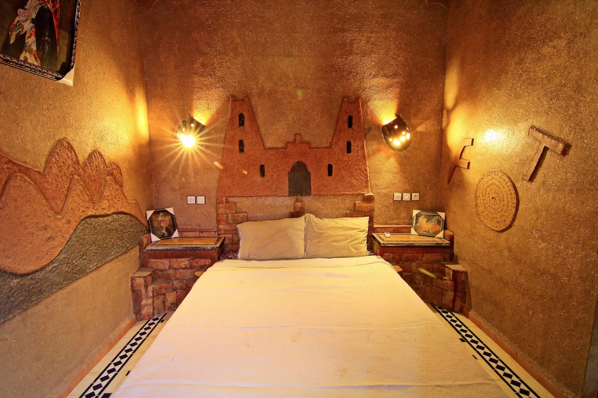 Una habitación con una cama, dos mesitas de noche, un mural en la pared y dos luces fijadas a la pared.