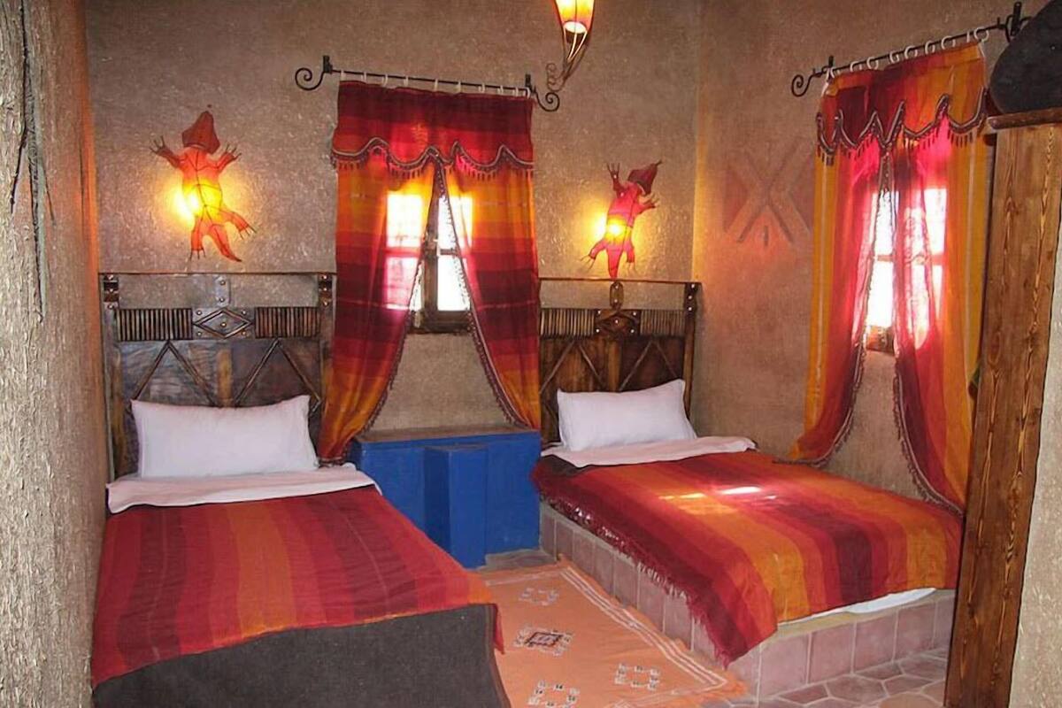 Habitación con dos camas, cada una con cabecero y una colcha rojo anaranjada. Se observan dos luces de pared con diseños de dragones.