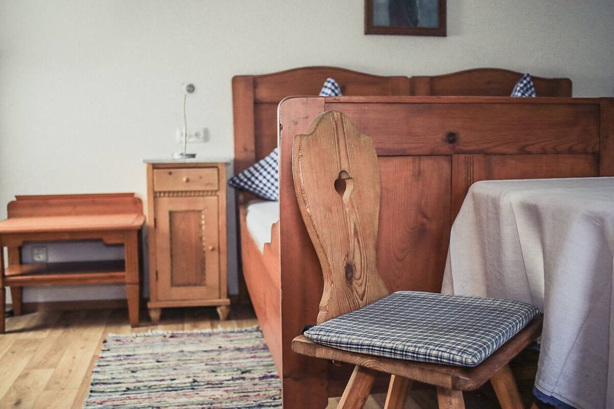 Un lit en bois avec une tête de lit, une table de chevet et un petit tabouret en bois surmonté d’un coussin à carreaux.