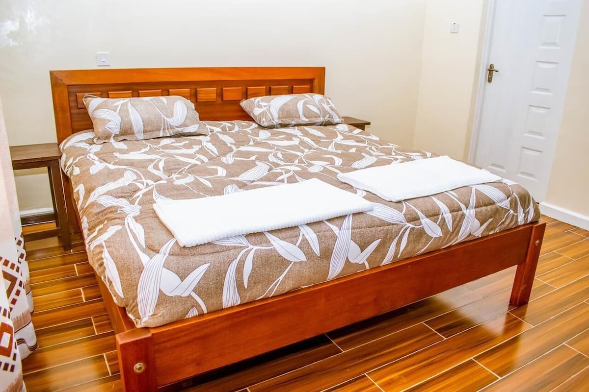 Un lit en bois avec une couvre-lit à motifs de feuilles et deux oreillers, une table de chevet et une porte blanche, le tout dans une pièce dont le sol est recouvert de parquet.