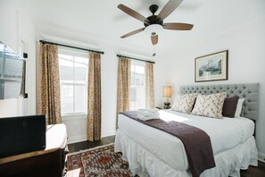 Une chambre à coucher avec un lit, un ventilateur de plafond, une fenêtre avec des rideaux, une table de chevet avec une lampe et un tableau encadré au mur.