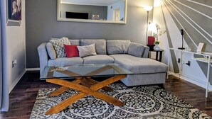 A modern living room with a grey sofa, a glass coffee table, a patterned rug, a large mirror, and a staircase with a railing. - Quiet Townhome in central Raleigh (Raleigh)