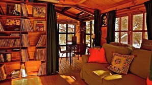 A cozy cabin interior with a wooden bookshelf filled with vinyl records, a comfortable sofa with a patterned cushion, and a dining area with a table and chairs. - A magical house in the heart of Guadarrama Park (Collado Hermoso)