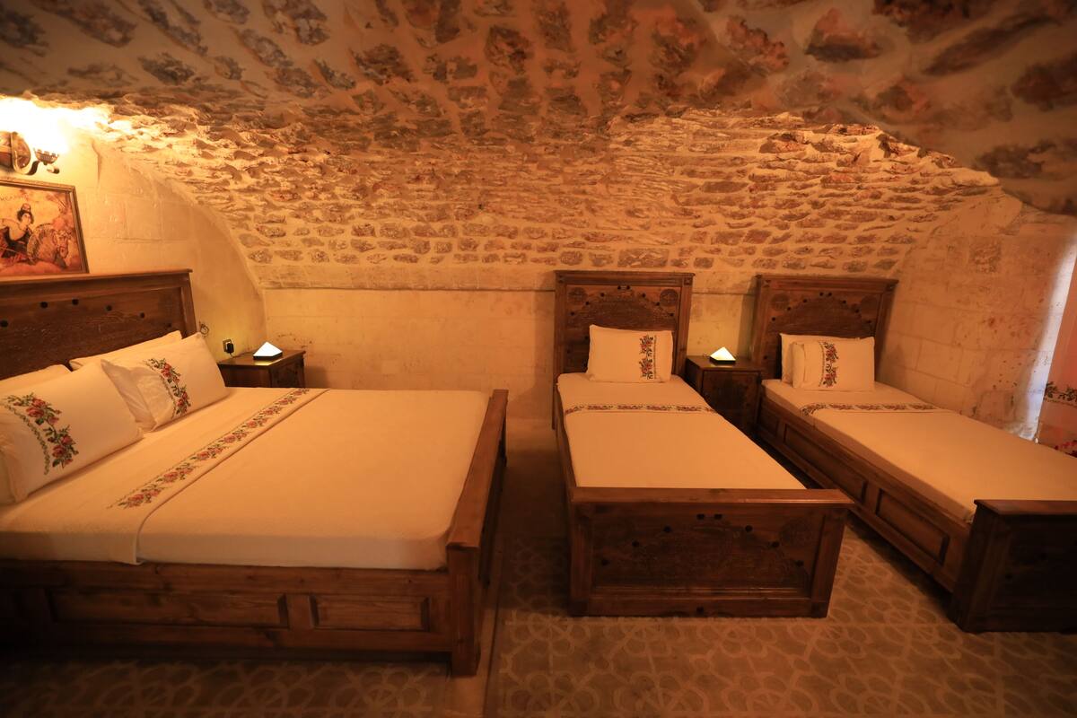 Ein Zimmer mit drei hölzernen Betten, jeweils mit Kopfteil und Nachttisch inklusive Lampe. Das Zimmer verfügt über eine Steindecke und -wände, sowie einen Teppich mit Muster.