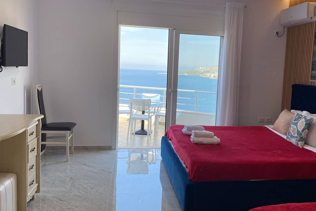 A bedroom with a bed, a desk, and a chair. There is a view of the ocean through a glass door.