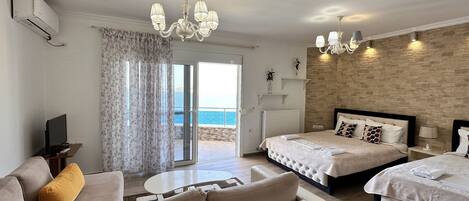 A hotel room with a sofa, two beds, a TV, a table, and a view of the sea.