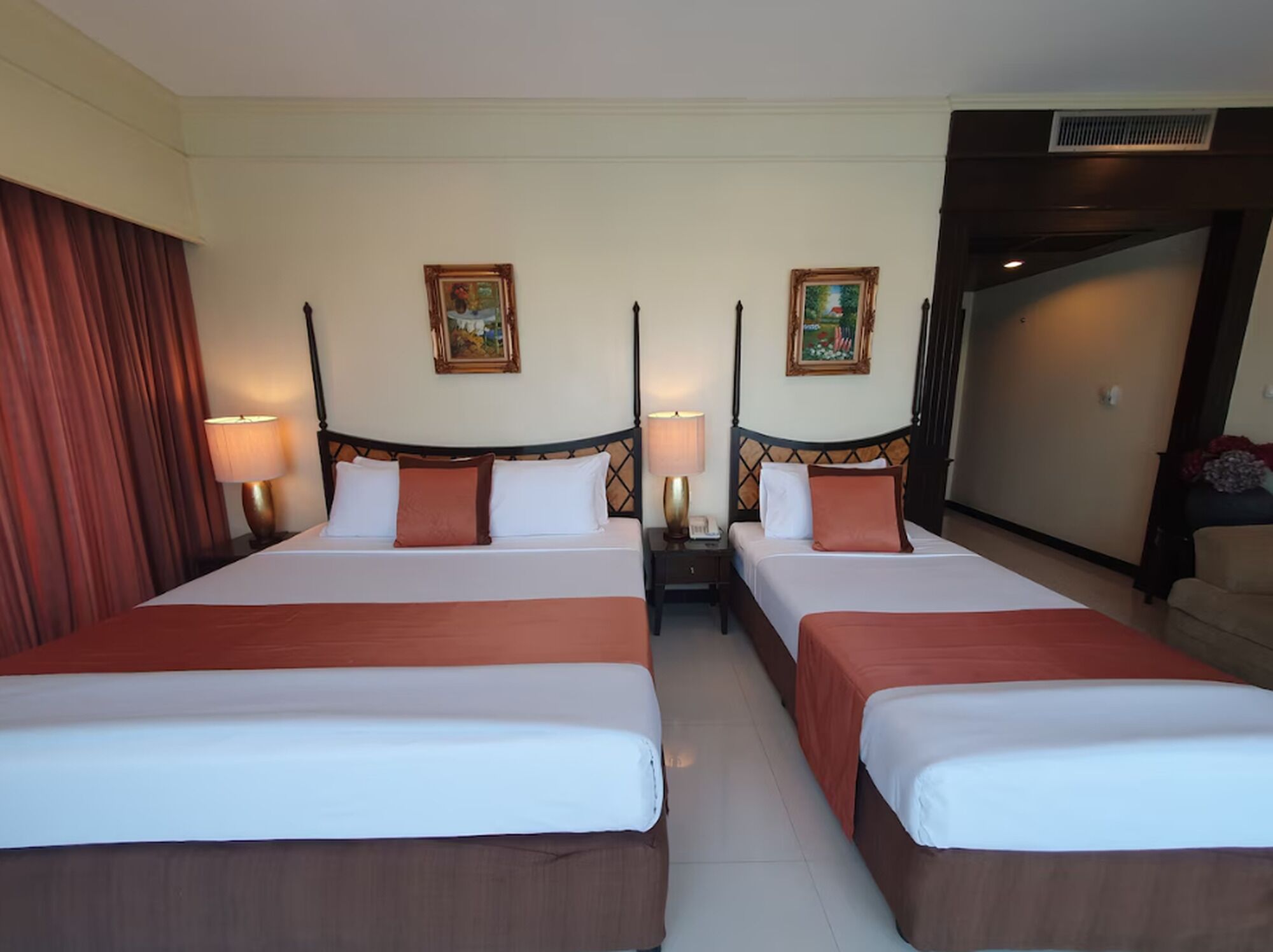 A hotel room with two beds, each with a wooden headboard, white bedding, and a red accent pillow. There are two bedside tables with lamps, a painting on the wall, and a door leading to another room.