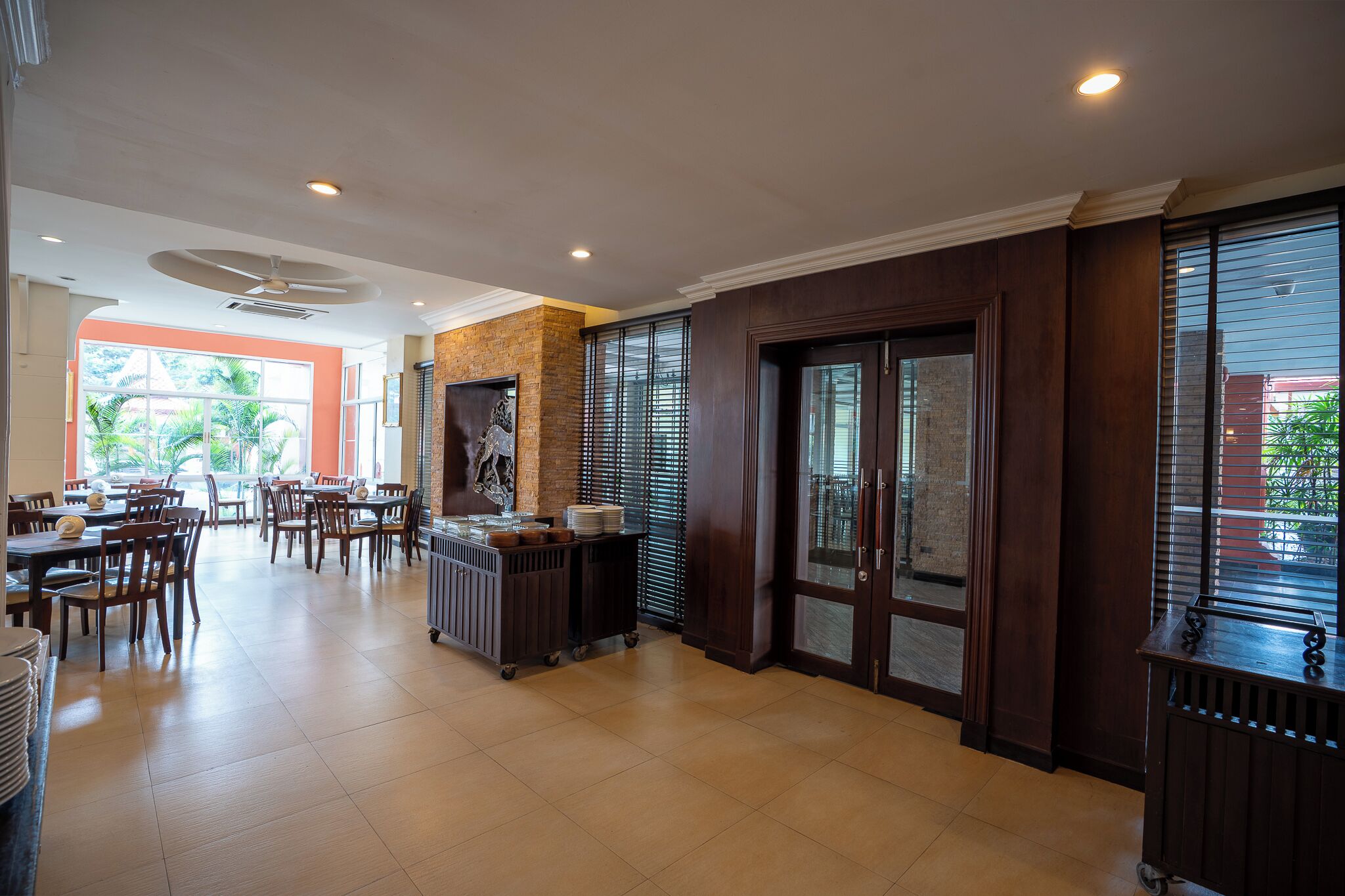 A spacious dining area with multiple tables and chairs, a central serving cart, and large windows with blinds.