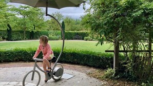 Une personne fait du vélo d’appartement sous un auvent, dans un jardin.