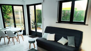 Un salon moderne avec un canapé noir, deux poufs gris, une table à manger avec des chaises et de grandes fenêtres donnant sur l’extérieur.
