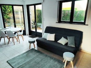 Una sala moderna con un sofá negro, dos otomanas grises, una mesa de comedor con sillas y ventanas amplias con vistas al exterior.