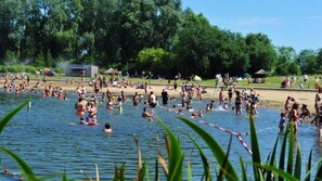 Un área de piscina al aire libre concurrida, con gente en el agua y en la orilla, rodeada de árboles.